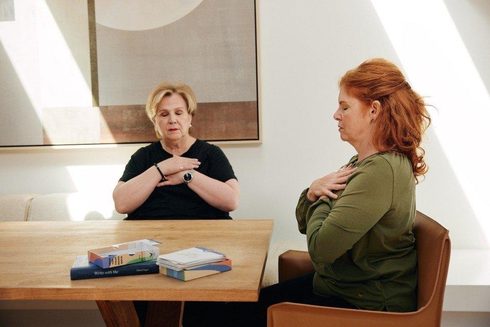 Two women seated at a table in a bright room, eyes closed and hands crossed over their chests, with a few books on the table beside them