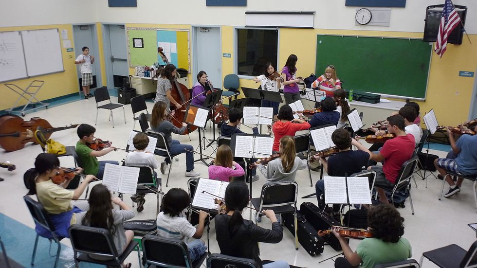 A full school orchestra rehearsing in a classroom, dozens of students at music stands playing violins, cellos and double basses while a teacher conducts from the front