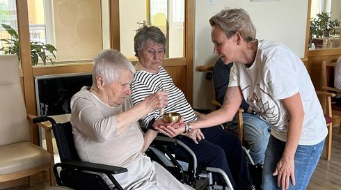 A woman steadying a small metal singing bowl in an older woman's hands while a second resident looks on, both seated in wheelchairs in a care home lounge