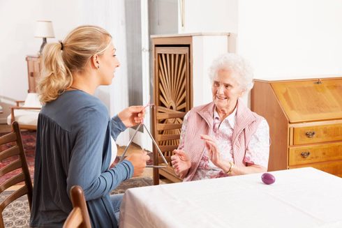 A woman holding a triangle beside an older woman who is clapping along at a dining table, with a shaker egg resting on the tablecloth